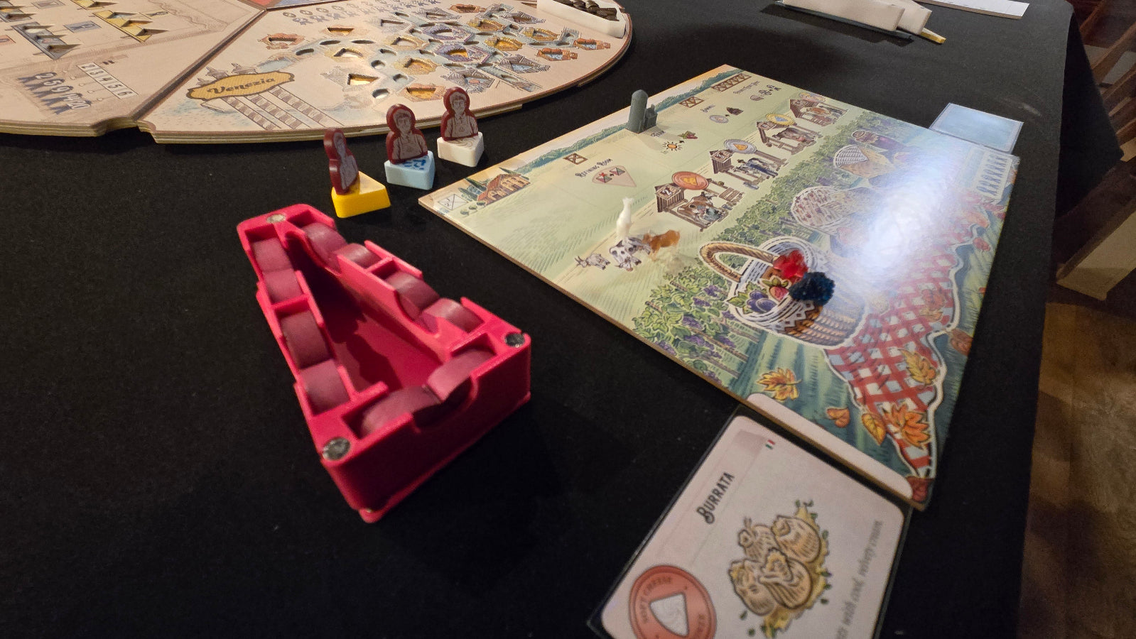 Board game components on a table with a colorful map and pink plastic piece.