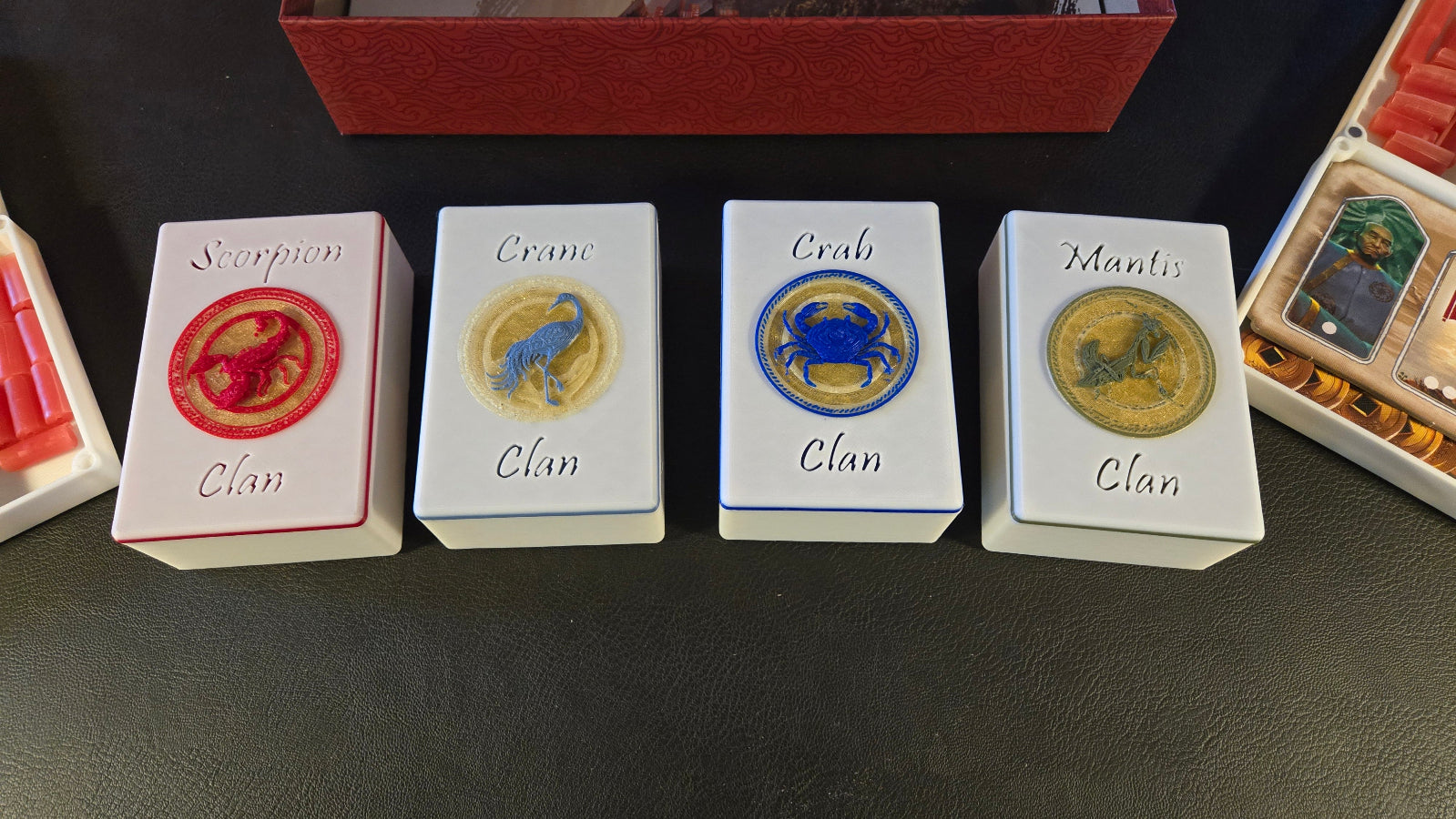 Four card packs labeled 'Scorpion Clan', 'Crane Clan', 'Crab Clan', and 'Mantis Clan' on a dark surface.