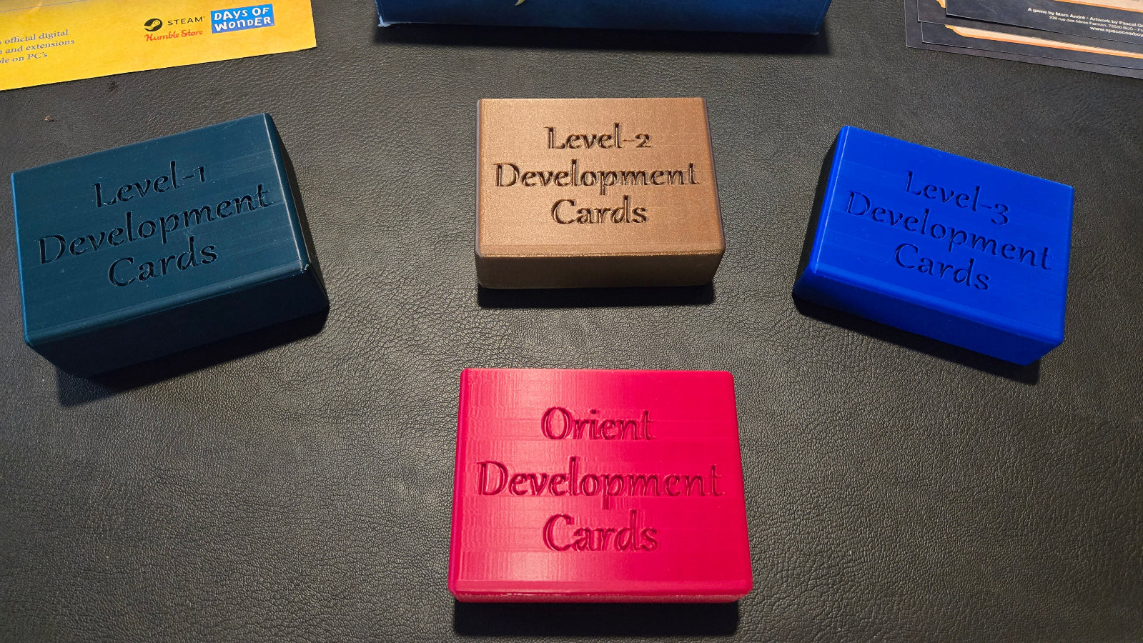 Four boxes labeled 'Level 1 Development Cards', 'Level 2 Development Cards', 'Level 3 Development Cards', and 'Orient Development Cards' on a dark surface.
